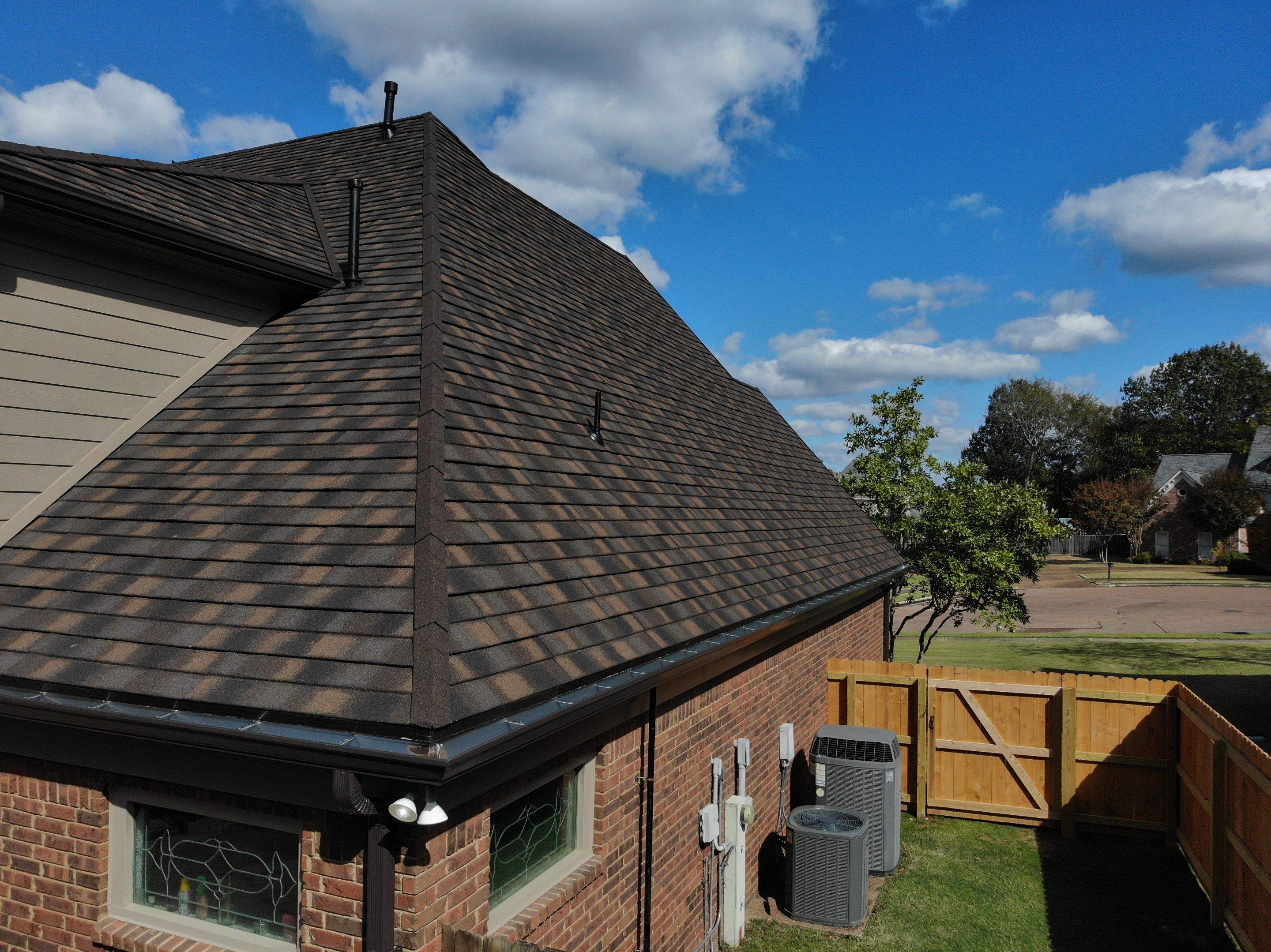 Dark brown shingle roof on a suburban brick home, featuring multiple roof slopes and vents under a bright blue sky with scattered clouds. A wooden fence and neighboring homes are visible in the background.